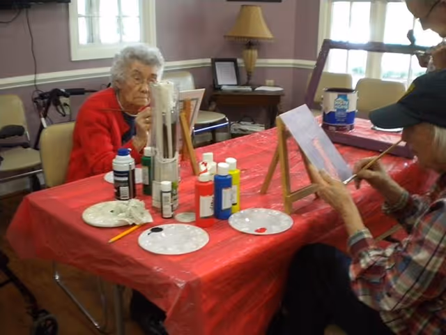 Two elderly individuals seated at a table covered with a red tablecloth, engaged in painting on canvases placed on small easels. Various paint bottles and palettes with paint are on the table. The room has a window, a lamp, and some furniture in the background.