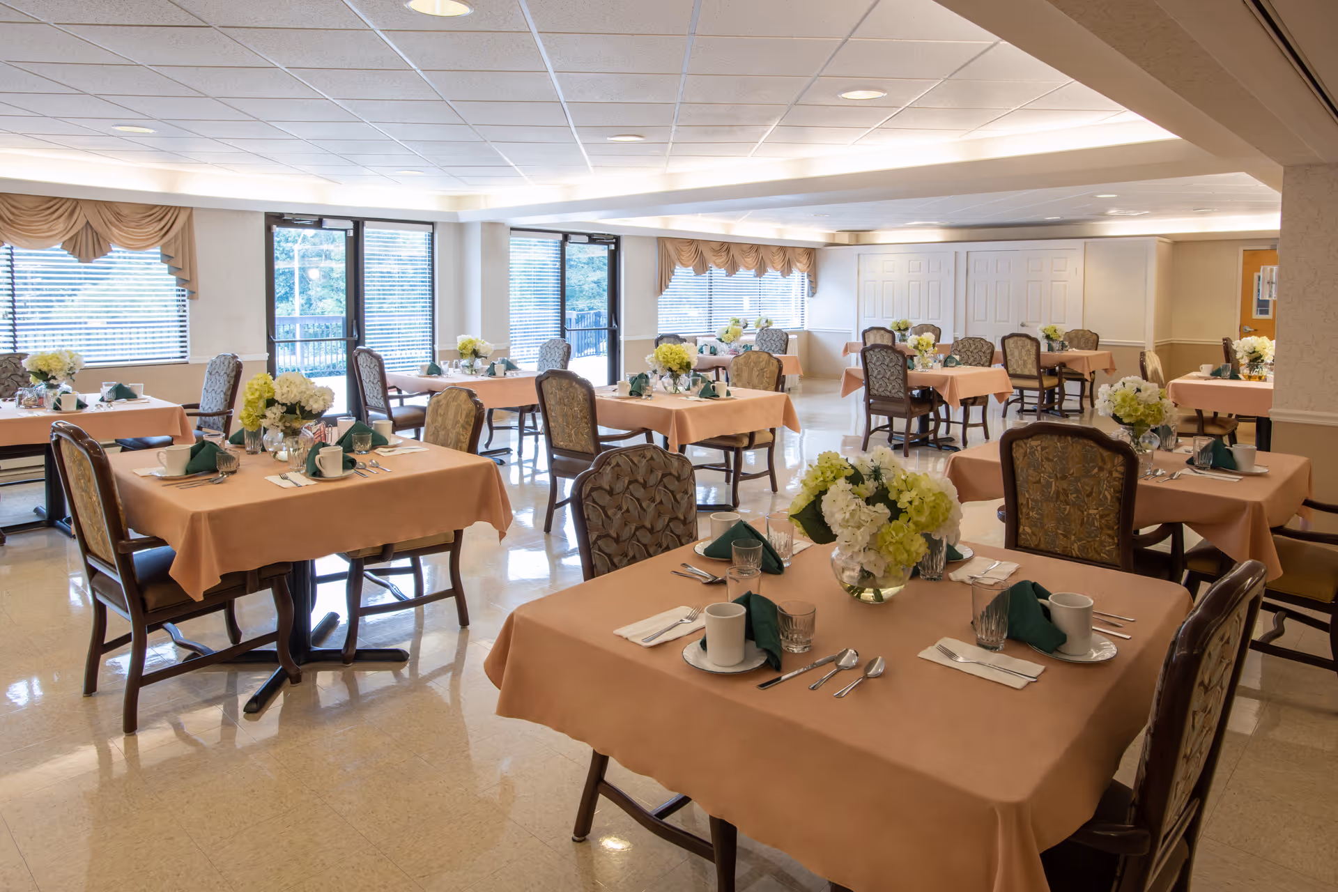 A dining room with several tables covered in light brown tablecloths, each set with white cups, glasses, silverware, and green folded napkins. Each table has a centerpiece of white and green flowers. The room has large windows with beige valances letting in natural light, and patterned upholstered chairs around the tables.