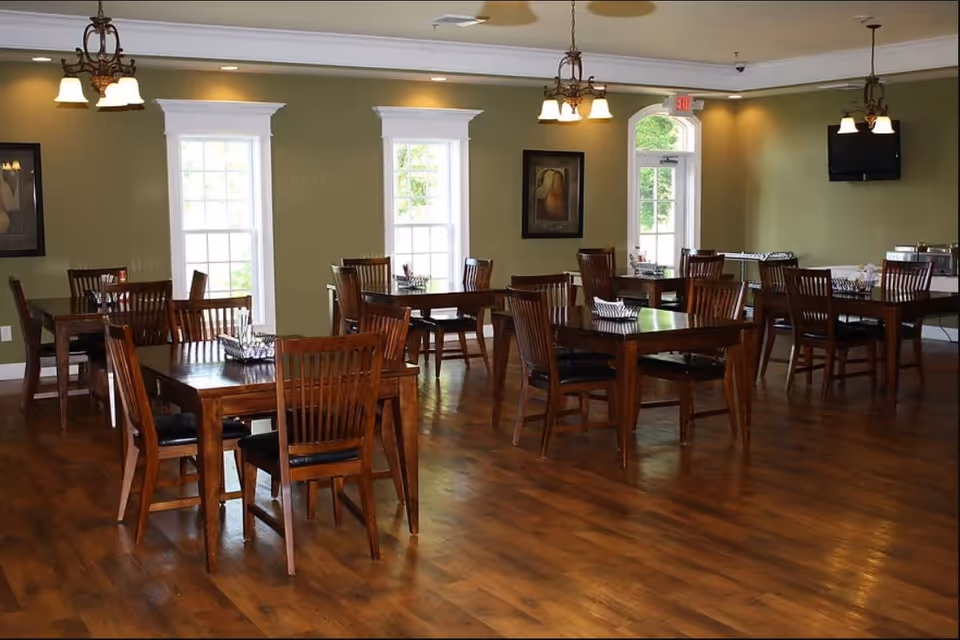 A dining room with multiple wooden tables and chairs arranged neatly on a polished wooden floor. The walls are painted green with white trim around the windows and door. There are three hanging light fixtures with multiple bulbs, framed artwork on the walls, and a television mounted on the far wall. Natural light comes through the windows and a door with an exit sign above it.