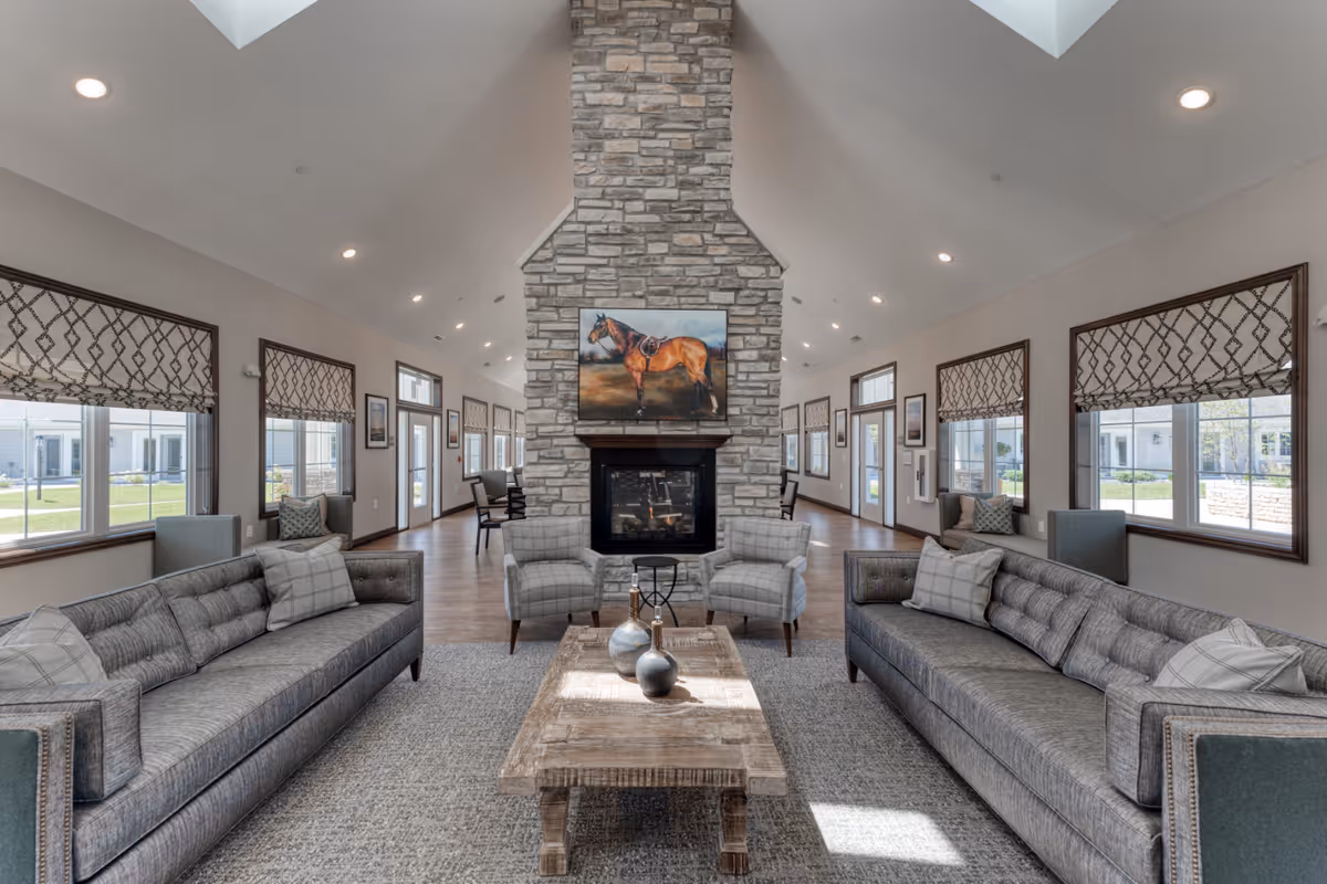Spacious senior living lounge with a central stone fireplace, two long sofas, armchairs, a wooden coffee table, and large windows under a vaulted ceiling.
