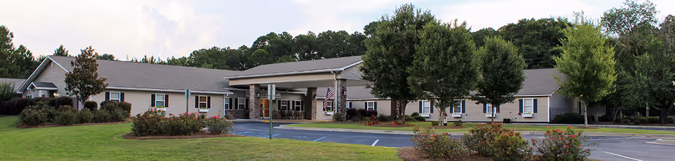 Exterior front view of a single-story senior living facility building with a covered entrance, surrounded by green lawns, trees, and bushes under a cloudy sky.