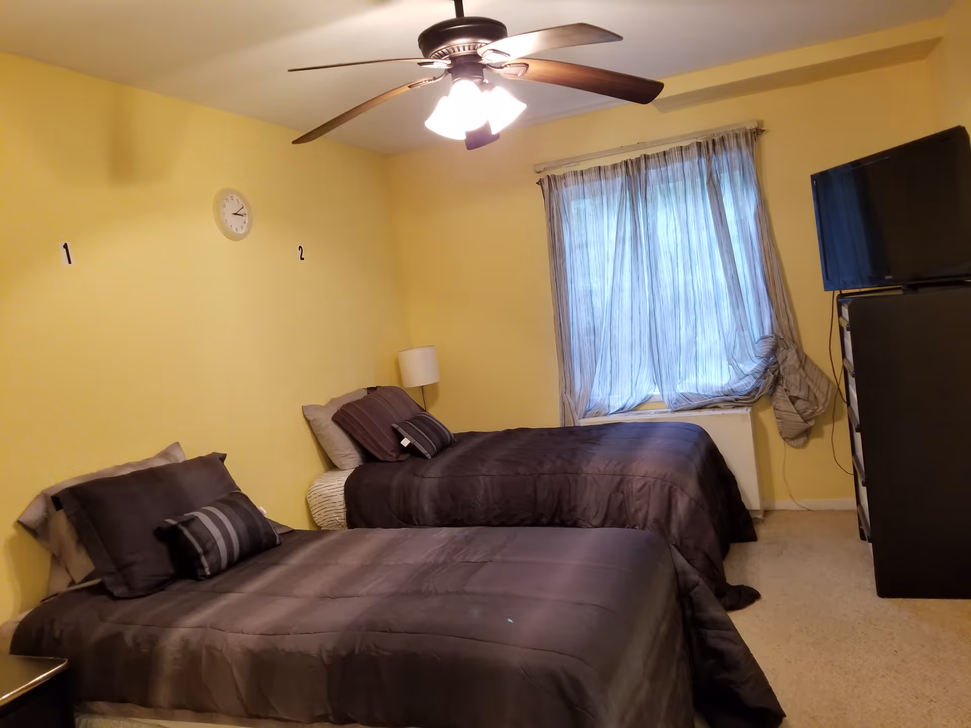 A bedroom with two single beds covered in dark gray bedding, each with matching pillows. The walls are painted yellow, and there is a white clock on the wall between the beds. A window with sheer curtains is on the far wall, and a ceiling fan with lights is mounted on the ceiling. A TV sits on top of a dresser to the right side of the room.