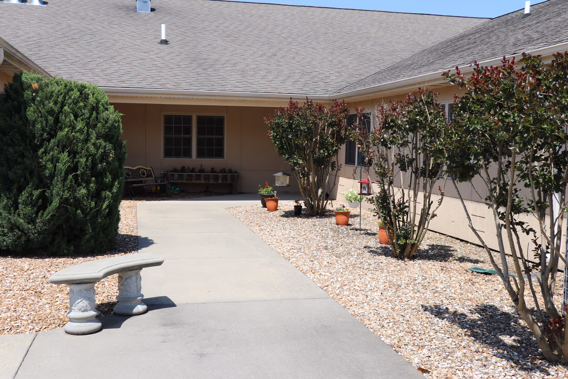 Concrete walkway leading to a healthcare facility courtyard with potted plants, shrubs, and a small stone bench.