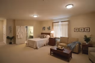 Spacious senior suite interior showing a bed, sofa, coffee table and bedside table with a lamp, plus a doorway to a bathroom.