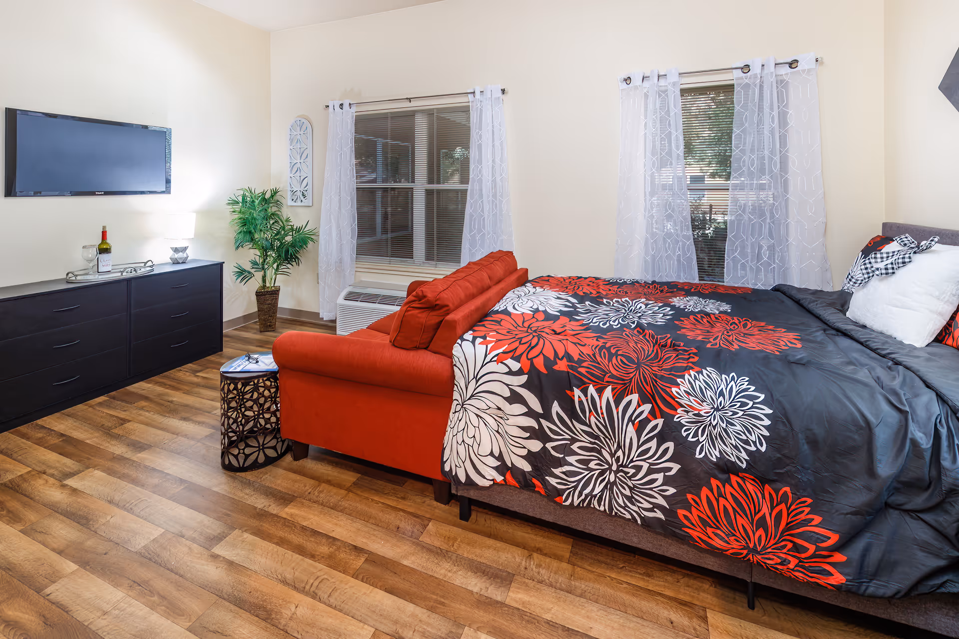 A bright bedroom with a large bed covered in a black, red, and white floral patterned comforter. Behind the bed is a red couch facing a black dresser with a flat-screen TV mounted on the wall above it. There is a small round side table next to the couch, a potted plant in the corner, and two windows with white sheer curtains allowing natural light into the room. The floor is wood with a warm tone.