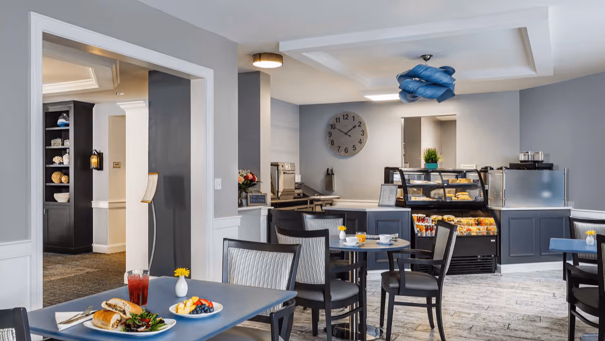 A dining area in a senior living facility with several tables and chairs. One table in the foreground has a plate with a sandwich, salad, and fruit, along with a glass of red beverage. In the background, there is a counter with a display case containing food items, a coffee machine, and a wall clock showing the time as 10:10. The room has light gray walls and a modern ceiling light fixture.