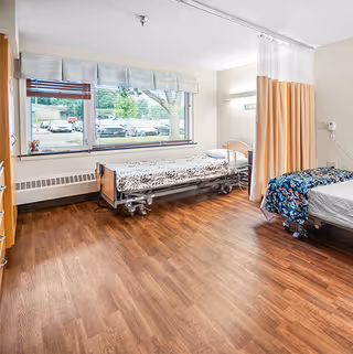 A bright room with two hospital-style beds, one near a large window with a view of parked cars and trees outside. The beds have patterned bedding, and a privacy curtain is partially drawn between them. The floor is wood, and the walls are light-colored.