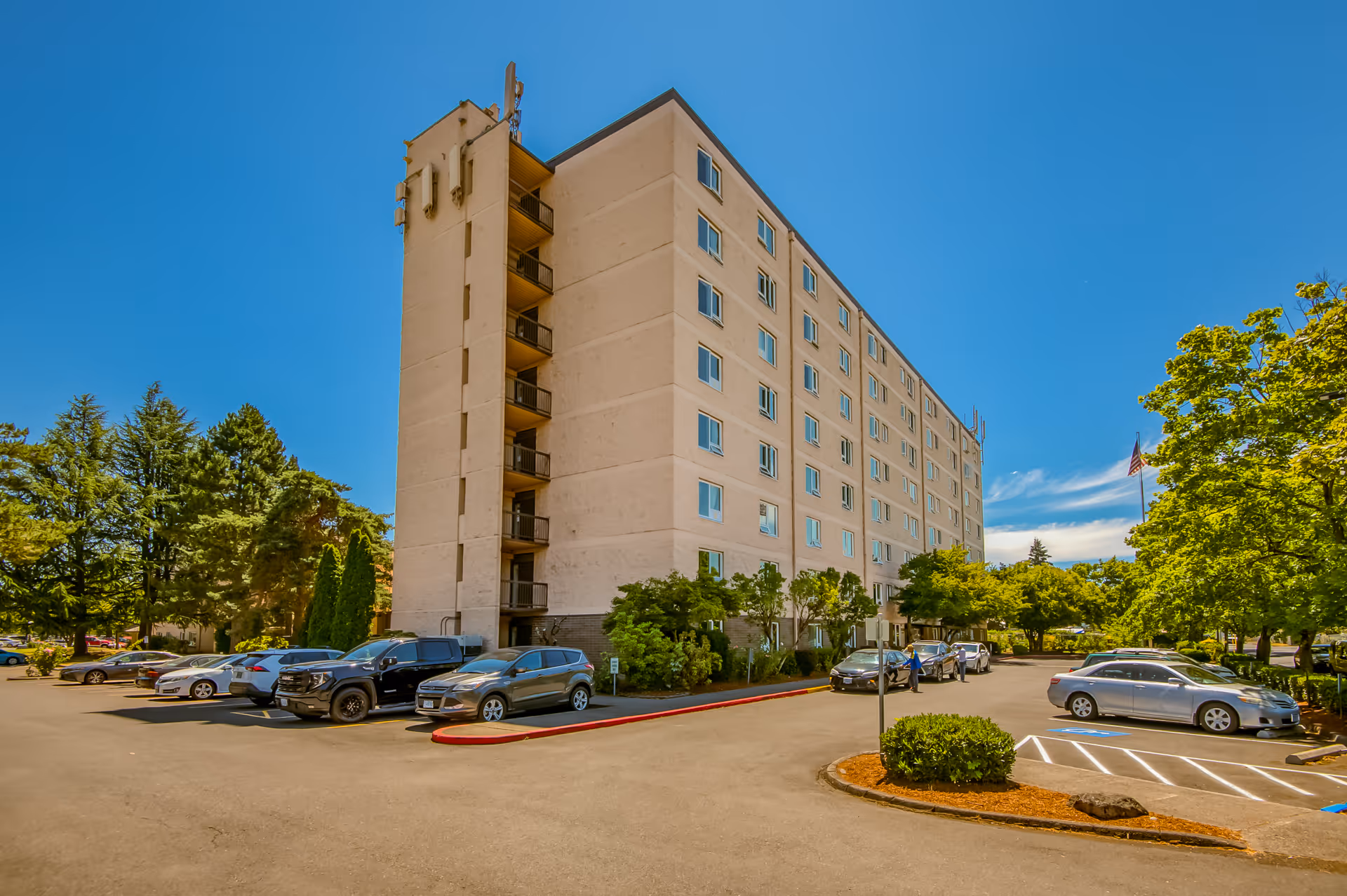 Exterior view of a multi-story senior living facility building named Kirkland Union Manors with a parking lot in front, several parked cars, trees, and a clear blue sky.