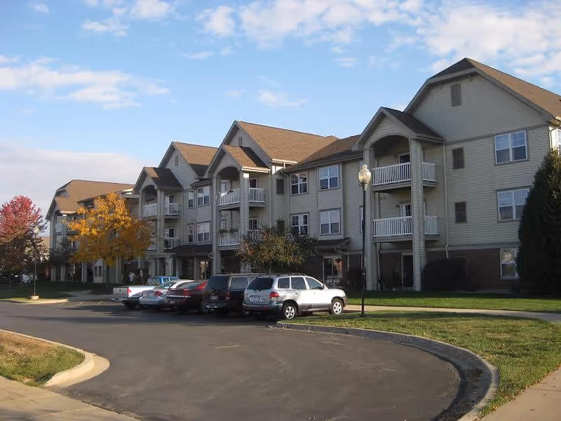 Exterior view of a three-story apartment building with beige siding and brown roofs, featuring balconies and multiple windows. Several cars are parked in front along a curved driveway, with green lawns and trees showing autumn colors under a partly cloudy sky.