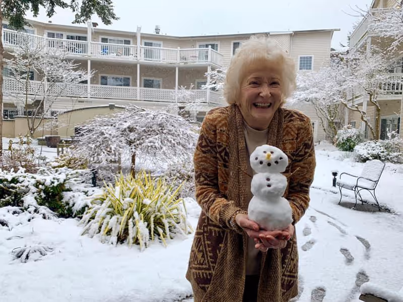 An elderly woman standing outside in a snowy courtyard holding a small snowman she made. She is smiling and wearing a patterned brown cardigan. The background shows snow-covered plants, benches, and a multi-story building with balconies.