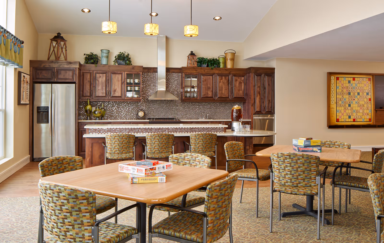 Communal dining and activity room with multiple tables and patterned chairs facing a full kitchen with wooden cabinets, pendant lights, and a refrigerator.