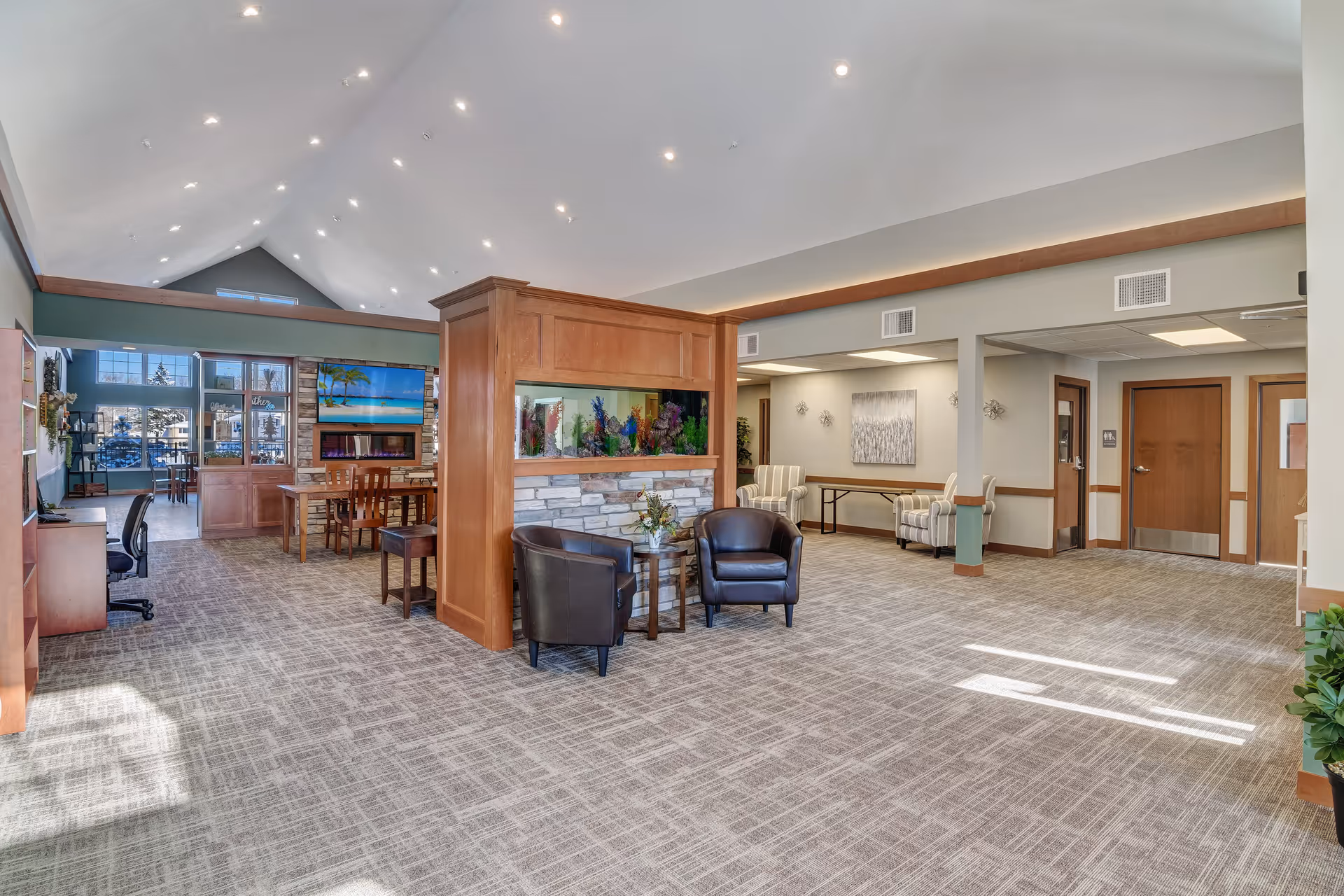 Spacious senior living facility common area with high vaulted ceiling and recessed lighting. The room features a large wooden partition with an aquarium and stone base, two black leather chairs with a small round table, and a seating area with two striped armchairs and a table. In the background, there is a dining area with wooden chairs and tables, a fireplace, and a large TV mounted above it. The space has carpeted floors and large windows letting in natural light.