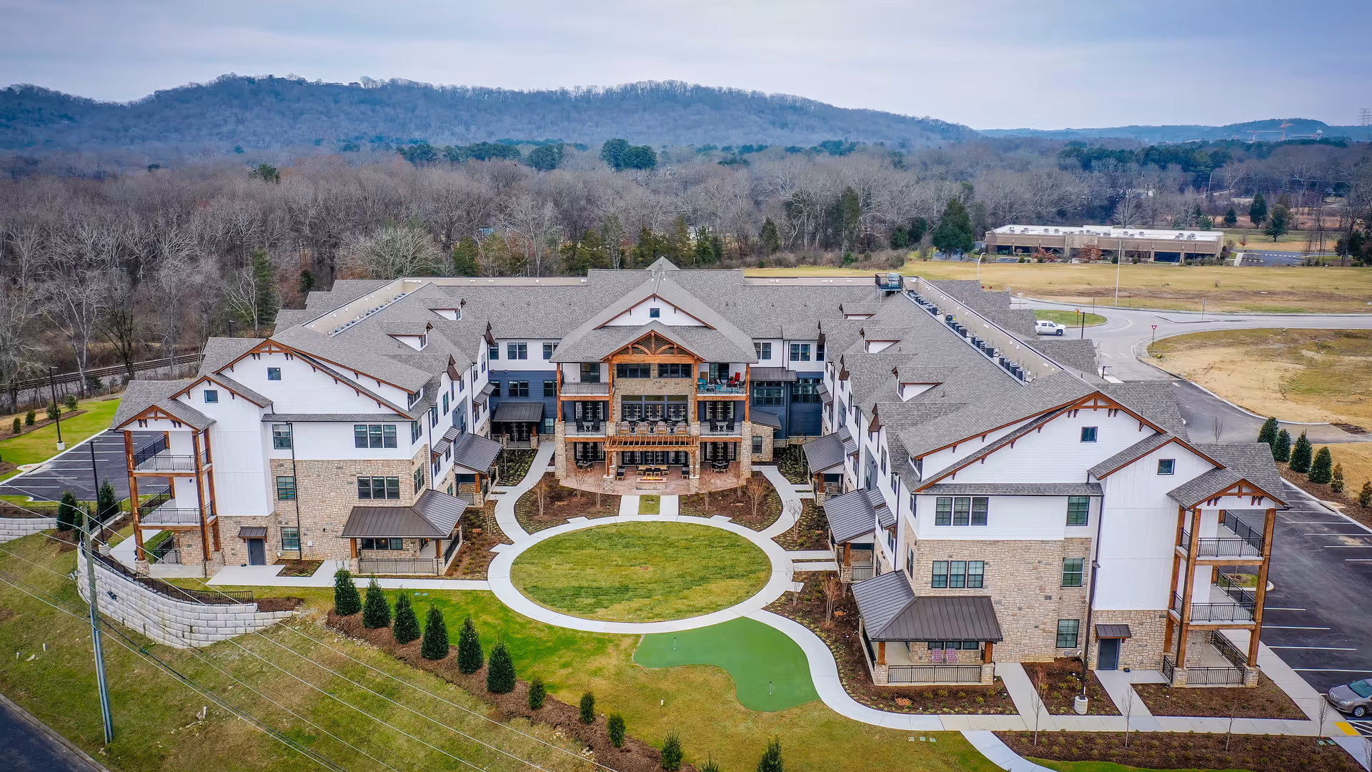 Aerial view of Everlan of Hixson, a large senior living facility with a U-shaped building surrounding a circular green lawn and walking paths. The building features multiple floors with balconies and a mix of stone and white siding exterior. The facility is set against a backdrop of wooded hills and open fields.