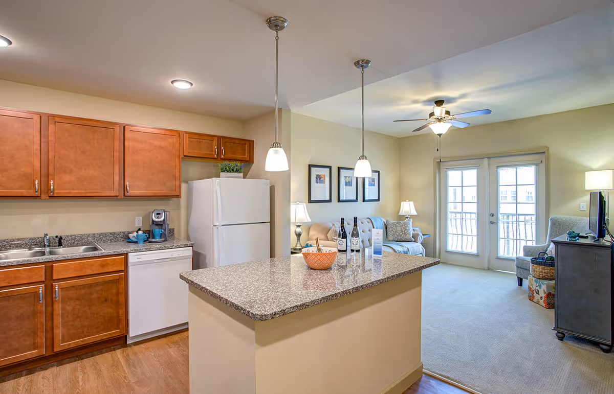 A bright and clean open-concept living space featuring a kitchen with wooden cabinets, granite countertops, a white refrigerator, dishwasher, and a coffee maker. The kitchen island has pendant lights hanging above it and holds a basket and wine bottles. Adjacent to the kitchen is a cozy living room with a sofa, armchair, lamps, framed pictures on the wall, a ceiling fan, and French doors leading to a balcony.