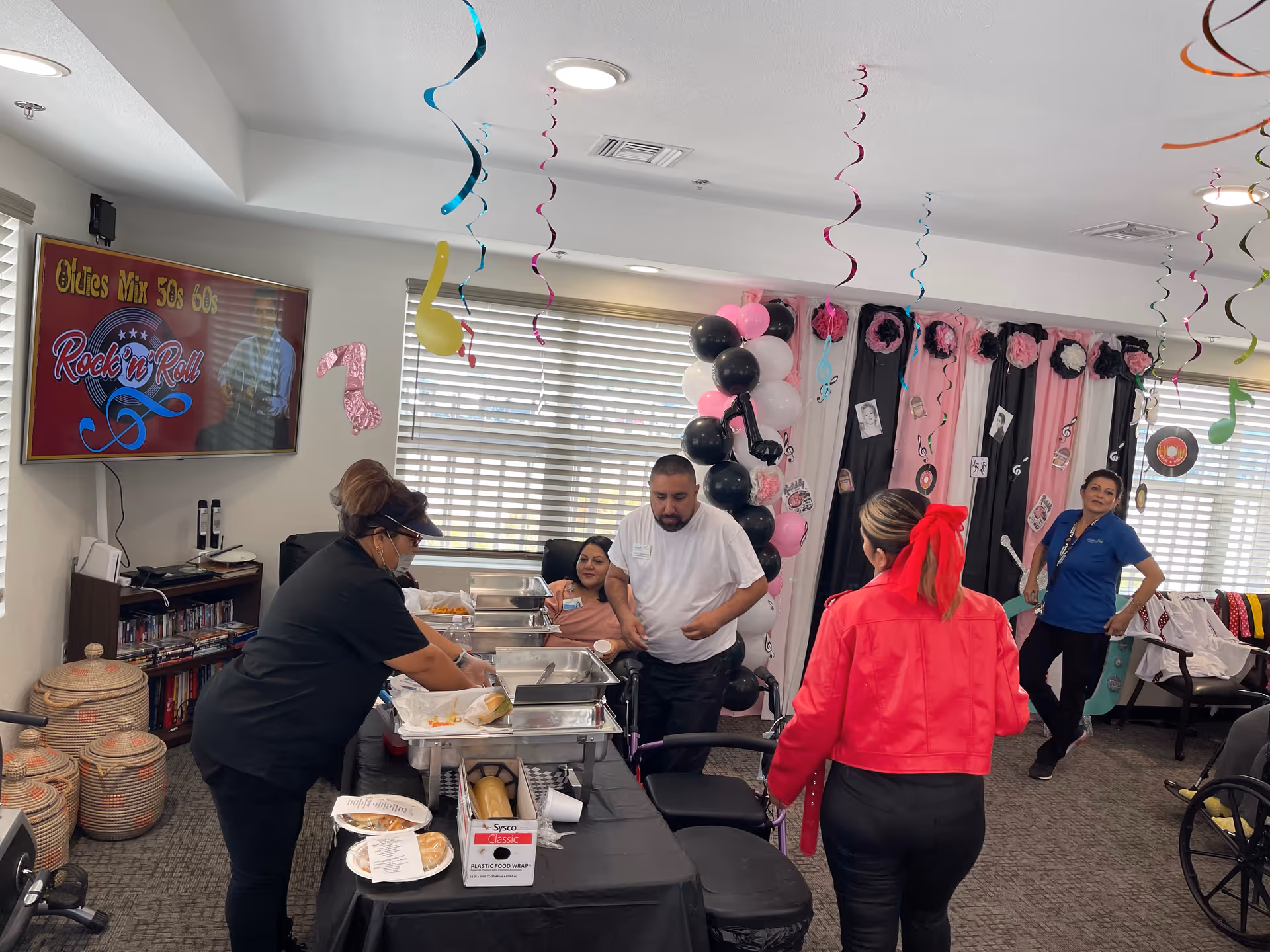 A decorated room with black, pink, and white balloons and hanging musical note decorations. Several people are present, including a woman serving food from chafing dishes on a table covered with a black tablecloth. A television screen on the wall displays 'Oldies Mix 50s 60s Rock n' Roll'. The room has large windows with blinds and various decorations on the walls.