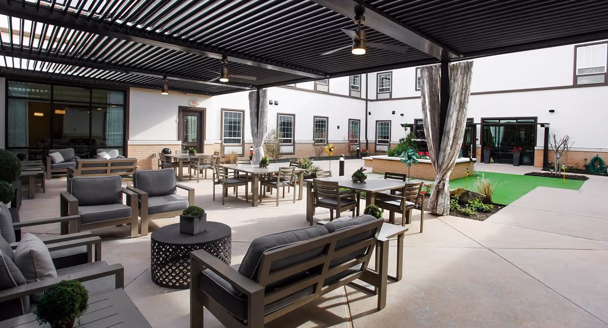 Outdoor courtyard area at a senior living facility with multiple seating arrangements including cushioned chairs and tables under a pergola, surrounded by a building with windows and doors. There is a small green putting area and some landscaping with plants and flowers.