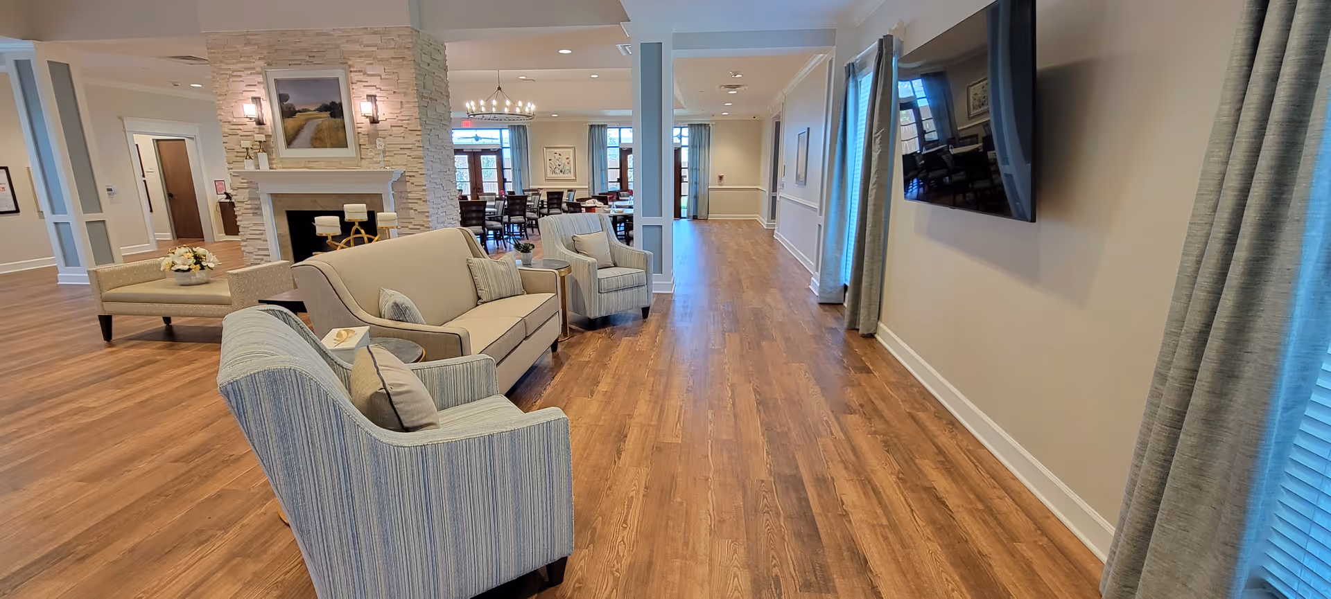 A spacious and well-lit living area in an assisted living facility featuring comfortable armchairs and a sofa arranged around a stone fireplace with a framed picture above it. The room has wooden flooring, a wall-mounted flat-screen TV, large windows with curtains, and a dining area visible in the background with a chandelier and multiple tables and chairs.