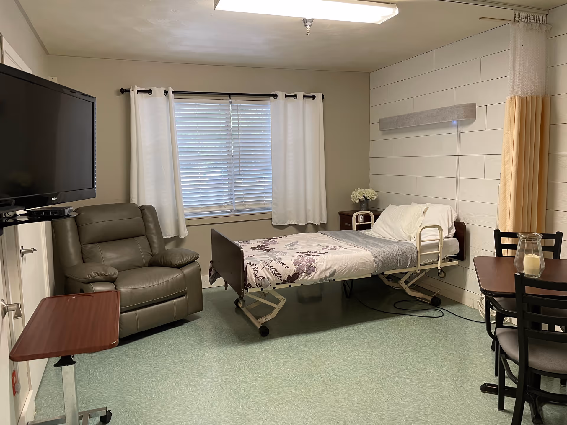 A simple patient room in a healthcare facility featuring a hospital bed with floral bedding, a gray recliner chair, a small wooden side table, a wall-mounted flat screen TV, a window with white curtains, a small dining table with four chairs, and a privacy curtain on the right side.