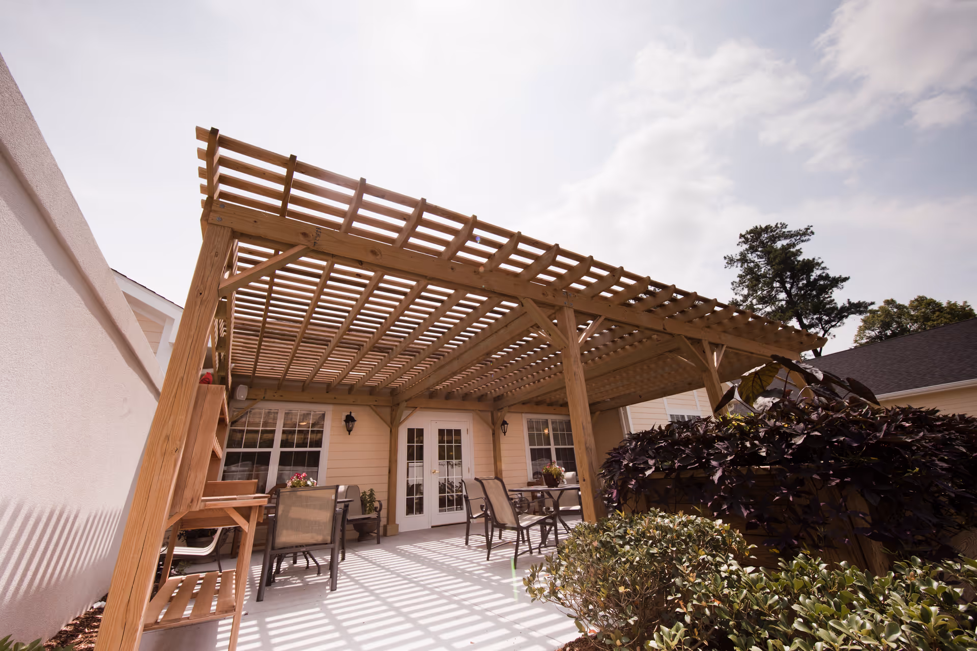 Outdoor patio area at TerraBella Hillsborough featuring a wooden pergola providing partial shade over a concrete floor. There are several tables and chairs arranged for seating, with potted plants and greenery around the perimeter. The building exterior has light-colored siding and white-framed windows and doors.