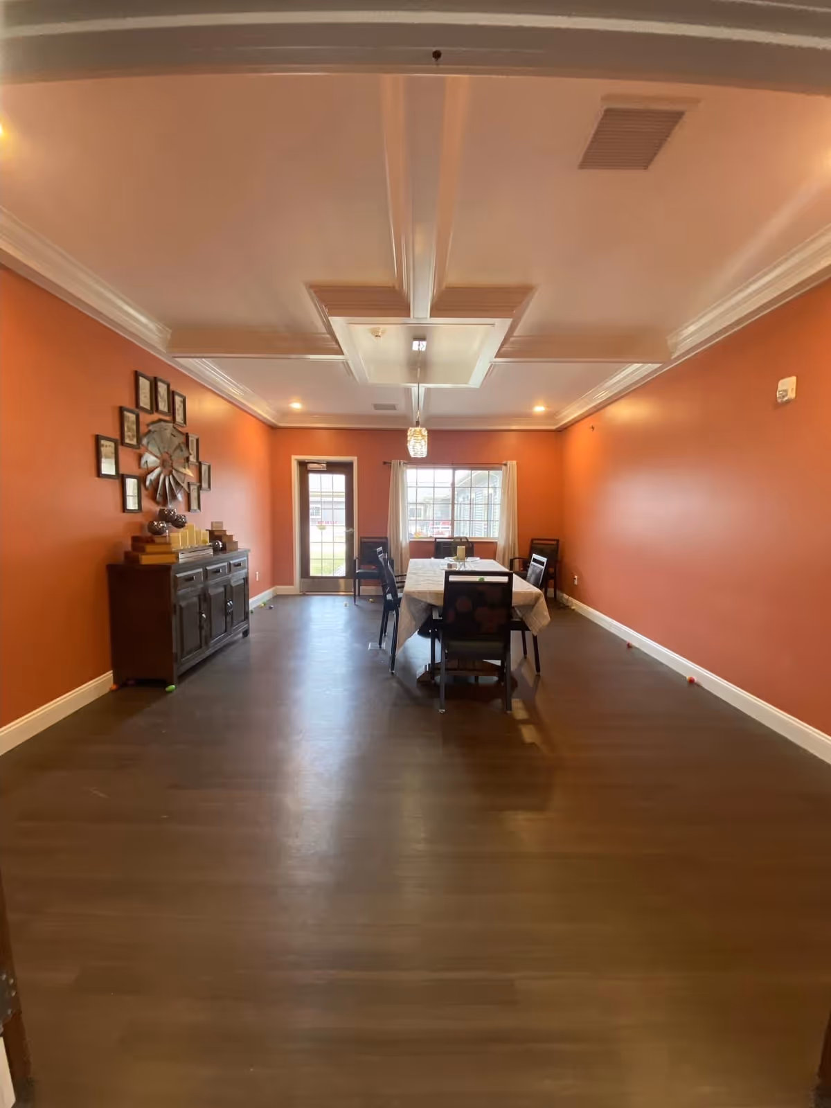 A dining room with orange walls, a wooden floor, and a white ceiling with decorative molding. There is a rectangular dining table covered with a tablecloth and surrounded by six chairs. A sideboard with decorative items and framed pictures is against the left wall. A door and a window with curtains are at the far end of the room.