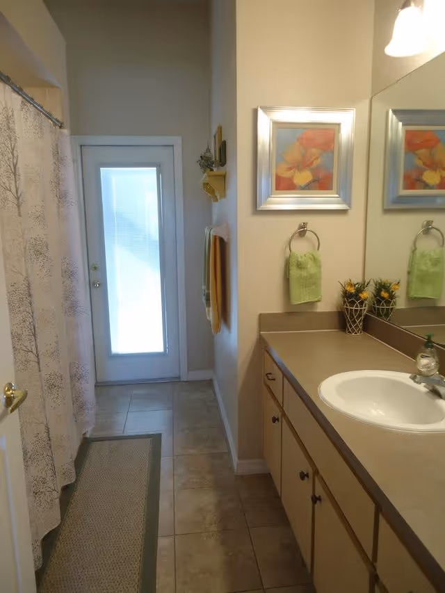 A bathroom with a tiled floor, a shower with a curtain featuring a tree pattern on the left, a long countertop with a sink on the right, a mirror above the sink, a green hand towel hanging on a ring, a small plant decoration on the counter, and a door with a frosted glass panel at the far end.