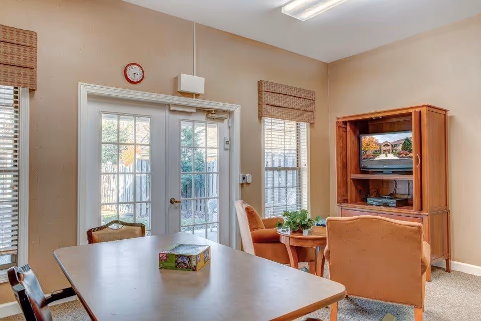 A cozy room with a rectangular table and chairs, a small round table with two armchairs, a wooden TV cabinet with a television displaying an outdoor scene, and French doors leading outside. The room has beige walls, windows with blinds, and a clock above the doors.