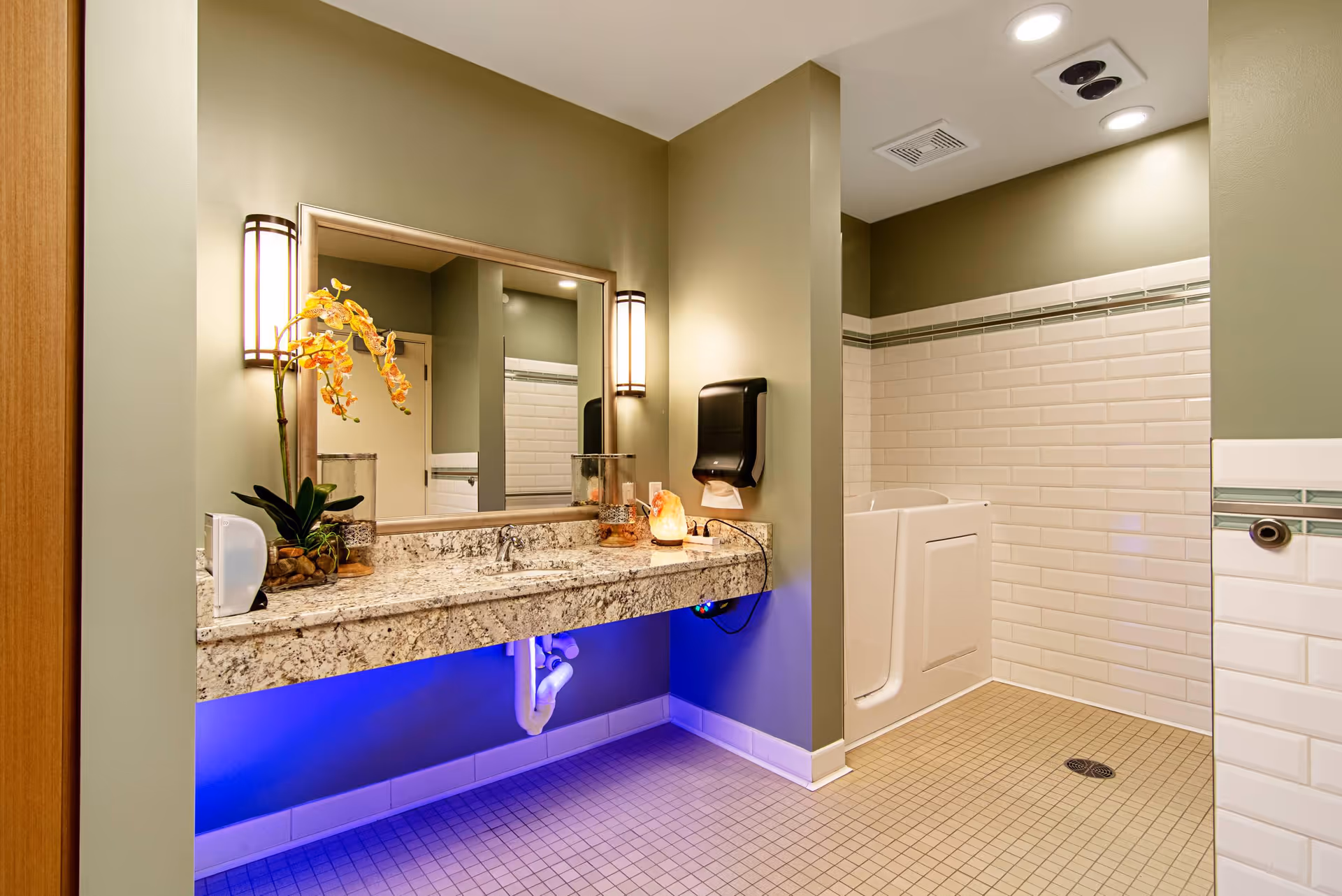 A clean and modern bathroom with a granite countertop sink, a large mirror with two wall-mounted lights on either side, a decorative orchid plant, and a walk-in shower with white subway tiles and a built-in seat. The floor is tiled with small square tiles, and there is blue accent lighting under the sink.