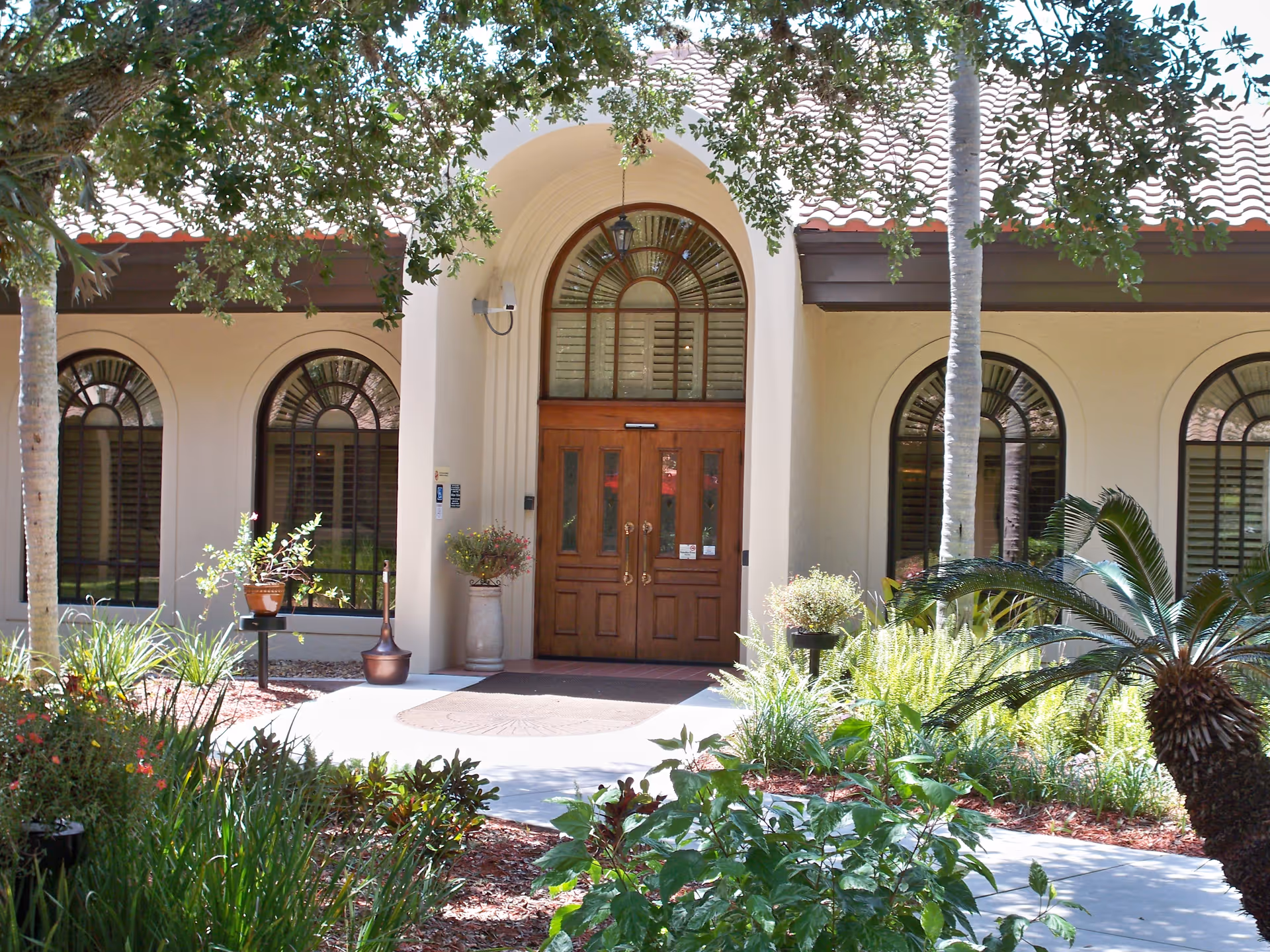 Entrance to a building with a wooden double door under an arched window. The building has cream-colored walls, arched windows with shutters, and a tiled roof. There are various green plants, shrubs, and trees in the landscaped garden area in front of the entrance.