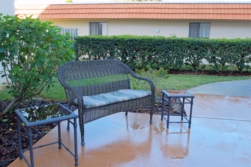 Outdoor patio area with a wicker bench that has a striped cushion, flanked by two small metal tables with glass tops. The ground is wet, reflecting the furniture. In the background, there is a hedge, some plants, and a building with a tiled roof and windows.
