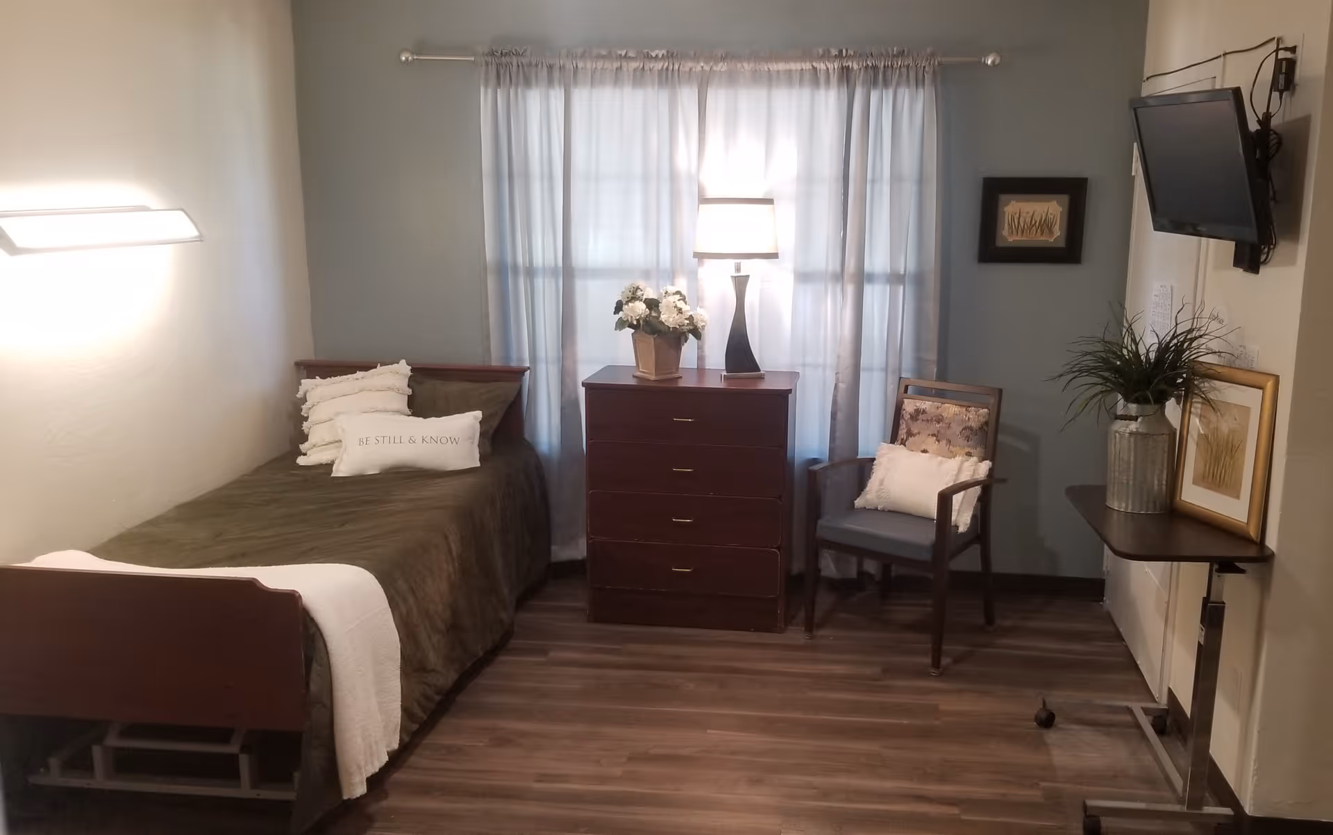 A small, cozy bedroom with a single bed covered in a dark green blanket and white pillows, one pillow has the text 'BE STILL & KNOW'. Next to the bed is a wooden dresser with a lamp and a flower pot on top. A window with sheer white curtains is behind the dresser. To the right, there is a wooden chair with a cushion and a small wall-mounted TV above a narrow table with a plant and framed artwork. The room has wooden flooring and soft lighting.
