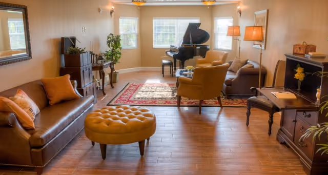 Spacious, well-lit living room with sofas, chairs, a round ottoman, an area rug, and a grand piano by three windows.