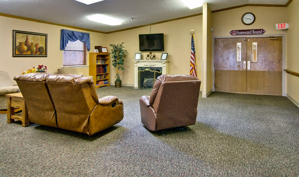 A cozy common area in a nursing center with two brown recliner chairs facing a fireplace and a wall-mounted TV. There is a bookshelf with books and framed photos, a potted plant, an American flag, and a window with blue curtains. Double wooden doors with a sign above reading 'Homeward Bound' and an exit sign are visible in the background.