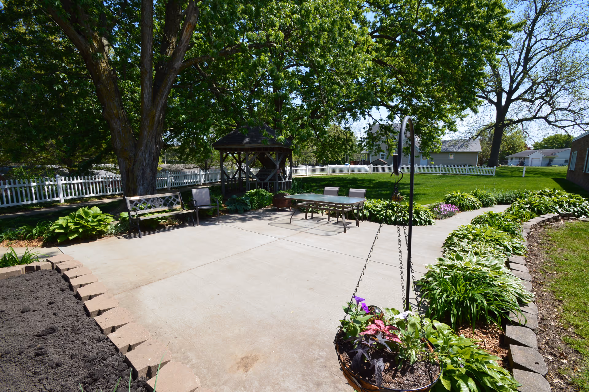 Sunny outdoor patio with seating, a gazebo, hanging planter, and surrounding greenery with a white picket fence.