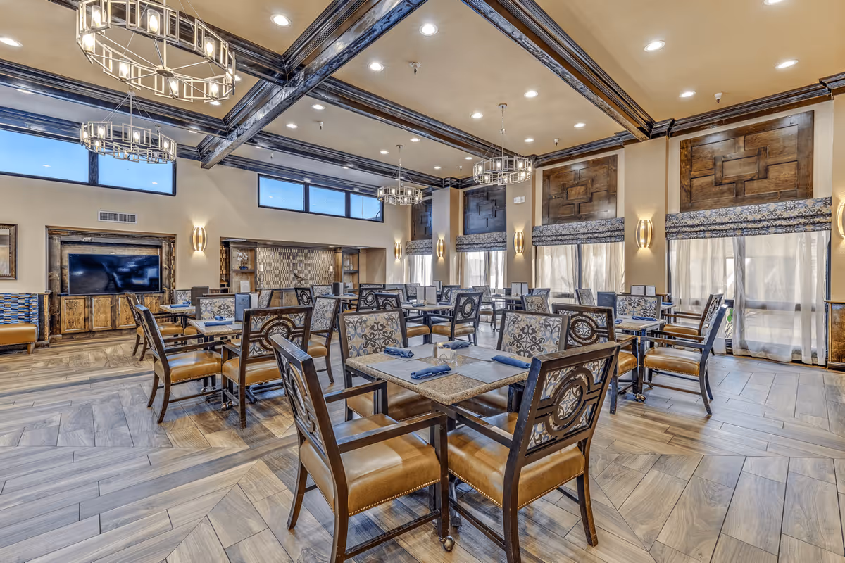 A spacious dining room with multiple tables and chairs arranged neatly. The room features large windows with decorative wooden panels and patterned curtains. Modern chandeliers hang from the ceiling, and there is a large flat-screen TV mounted on a wooden cabinet against one wall. The floor has a wood-like tile pattern, and the overall decor is warm and inviting.