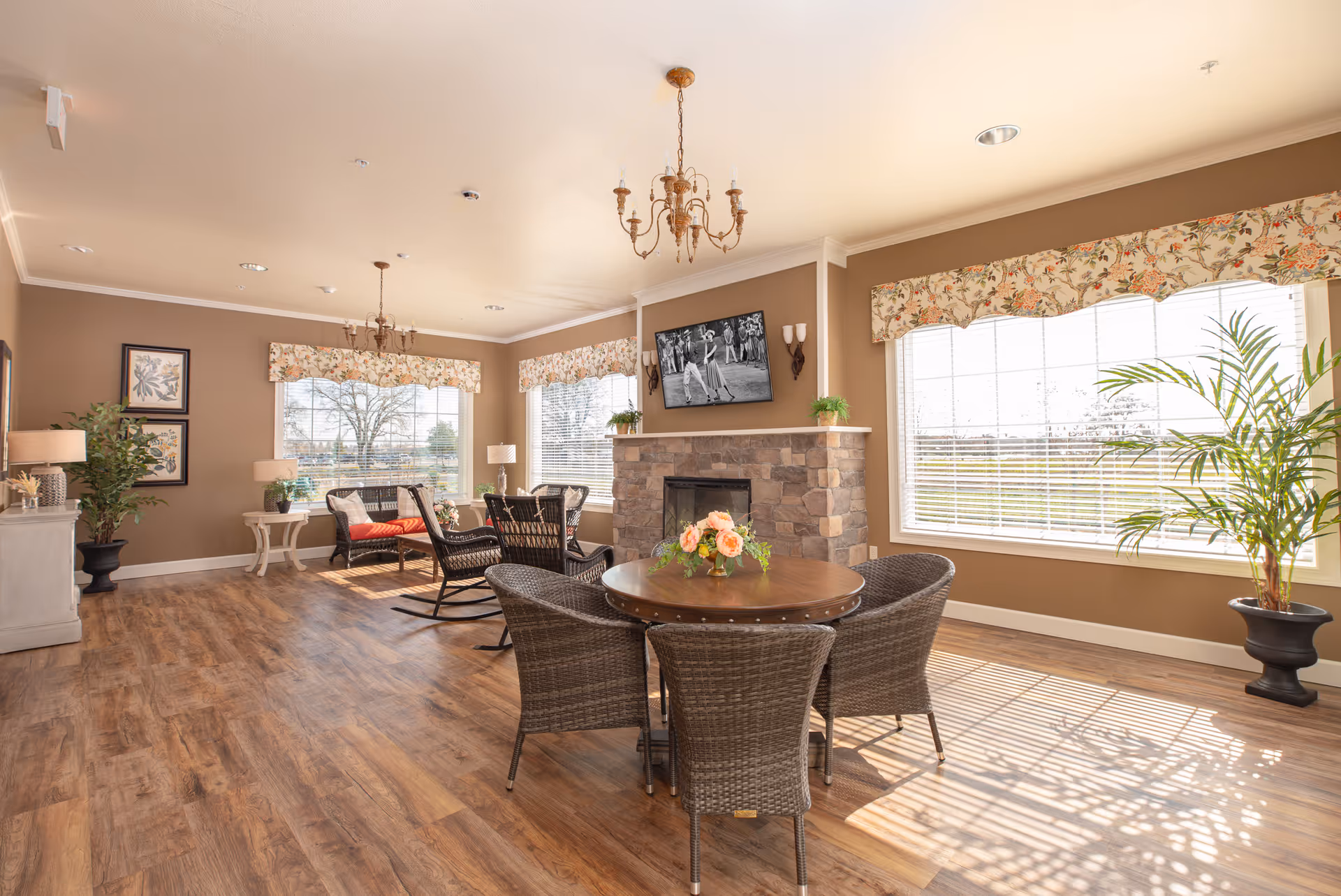 A bright and spacious living room with large windows letting in natural light. The room features a round wooden table with four wicker chairs, a stone fireplace with a flat-screen TV mounted above it, and two chandeliers hanging from the ceiling. There are floral valances on the windows, potted plants, and comfortable seating including a bench with cushions and rocking chairs. The floor is wood, and the walls are painted a warm beige color.