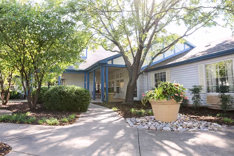 Exterior view of a senior living facility entrance with a concrete pathway leading to a covered porch. The building has white siding with blue trim, large windows, and is surrounded by trees, bushes, and a large planter with greenery and flowers.