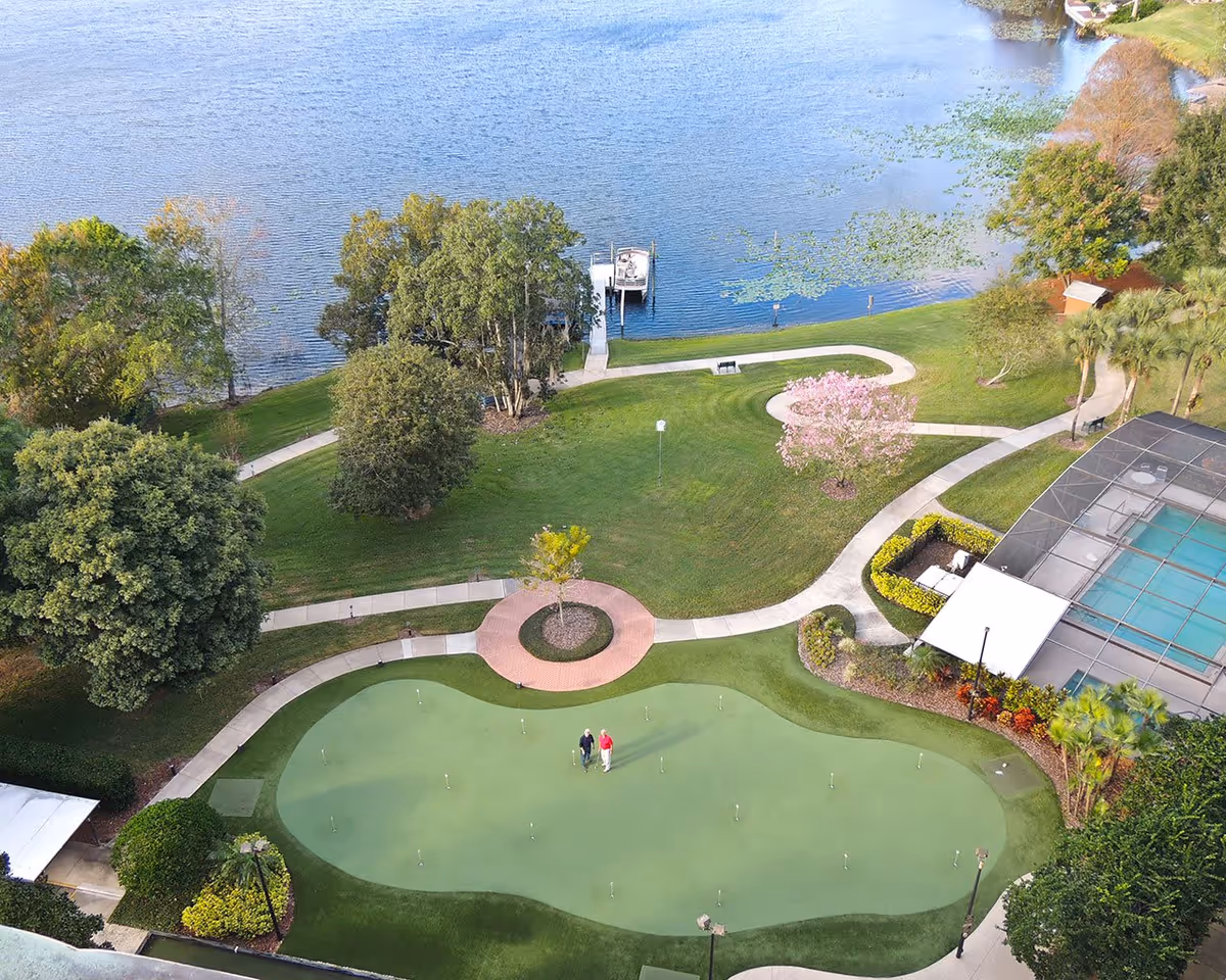 Aerial view of a landscaped outdoor area at Westminster Winter Park featuring a putting green with two people standing on it, winding walking paths, various trees including a flowering tree, a dock extending into a lake, and a screened-in swimming pool area.
