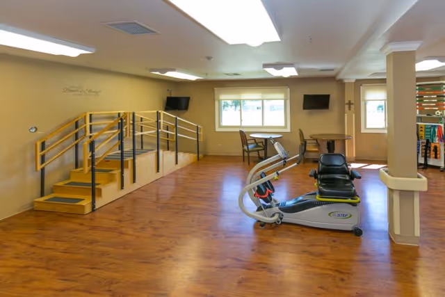 Spacious rehabilitation/exercise room with a recumbent stepper, practice stairs with handrails, tables and chairs, and wood-look flooring.