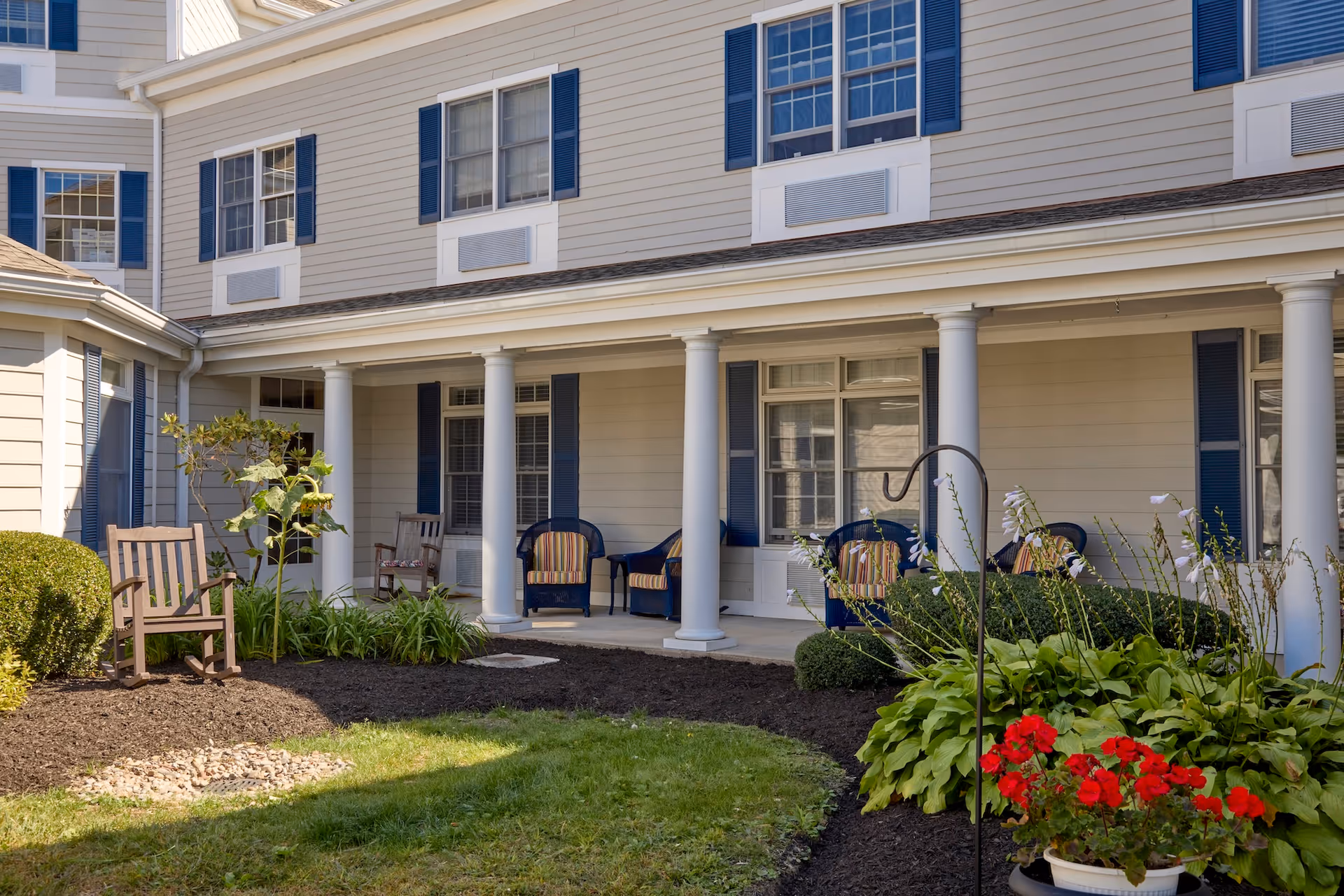Outdoor patio area of a senior living facility with white columns, beige siding, blue shutters, and several chairs with striped cushions arranged along the porch. The foreground features a garden with green plants, red flowers, and a grassy lawn.