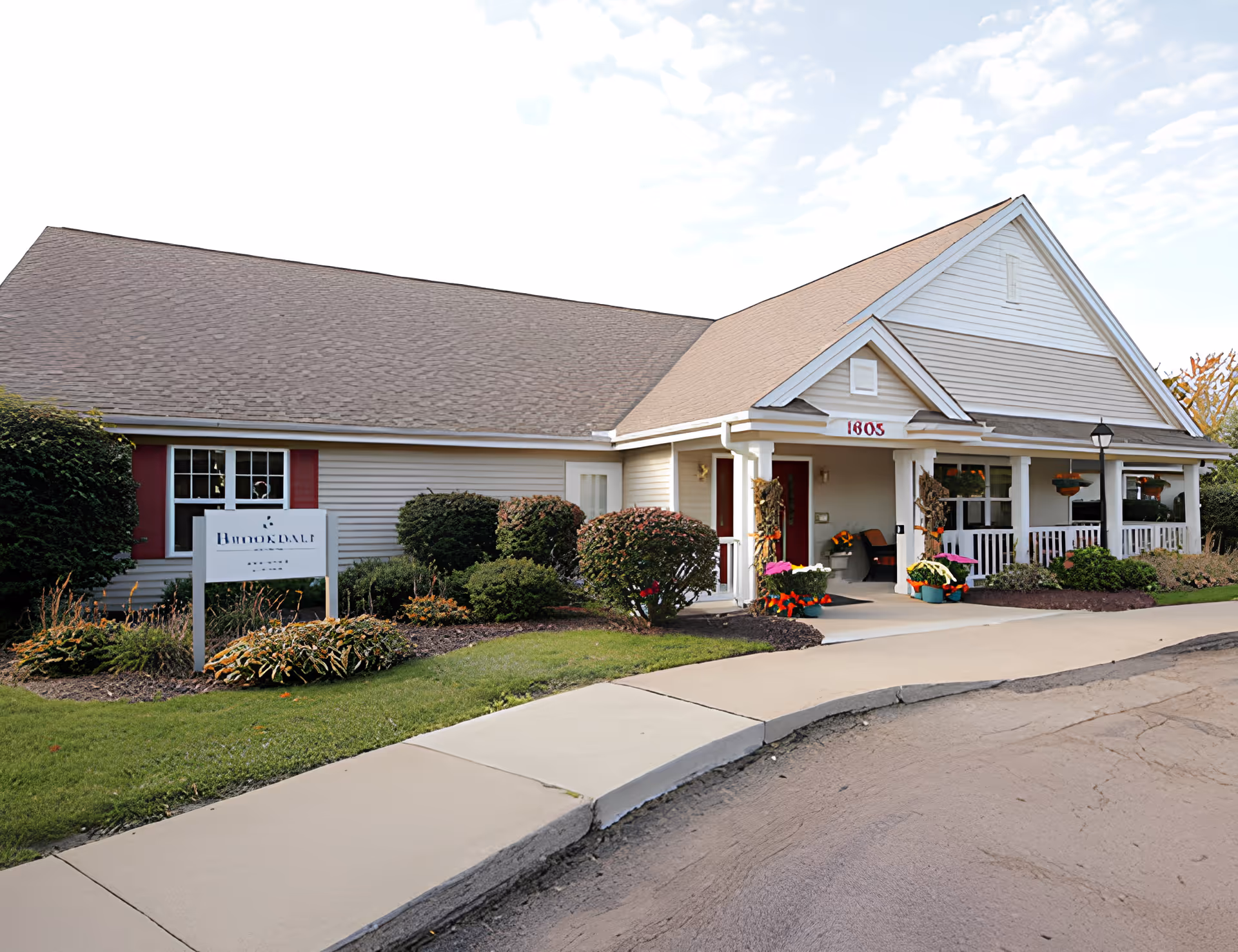 Exterior view of a single-story building with beige siding and a brown shingled roof. The building has a covered porch with white railings and columns, decorated with potted flowers and plants. There is a sidewalk and a well-maintained lawn with shrubs and bushes in front. A sign near the building reads 'Brookdale Monroe'.