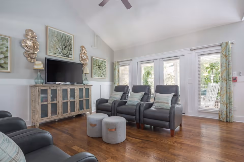 A bright living room with three black leather armchairs each with a decorative pillow, two round gray ottomans, a rustic wooden cabinet with a flat-screen TV on top, and large windows with floral curtains letting in natural light.