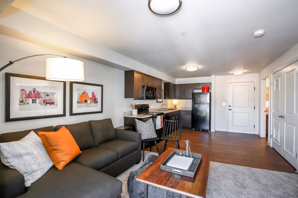 Open-plan living area with a dark sectional sofa and coffee table facing a kitchenette with dark cabinets and a black refrigerator.