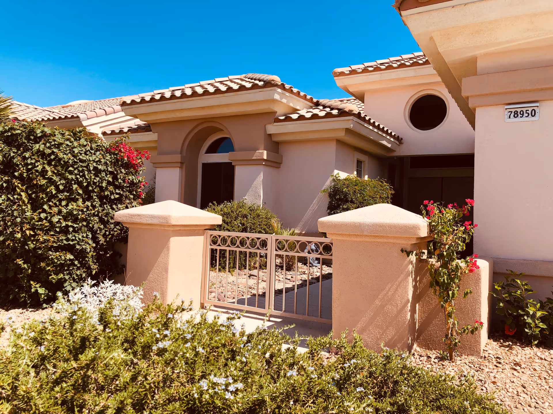 Exterior view of a single-story residential building with beige stucco walls and a tiled roof under a clear blue sky. The entrance features a small gated courtyard with plants and bushes surrounding the area.