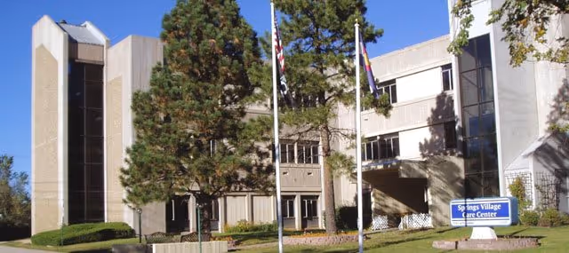 Front exterior of Springs Village Care Center showing the multi-story building, flagpoles, trees, and a lawn sign.