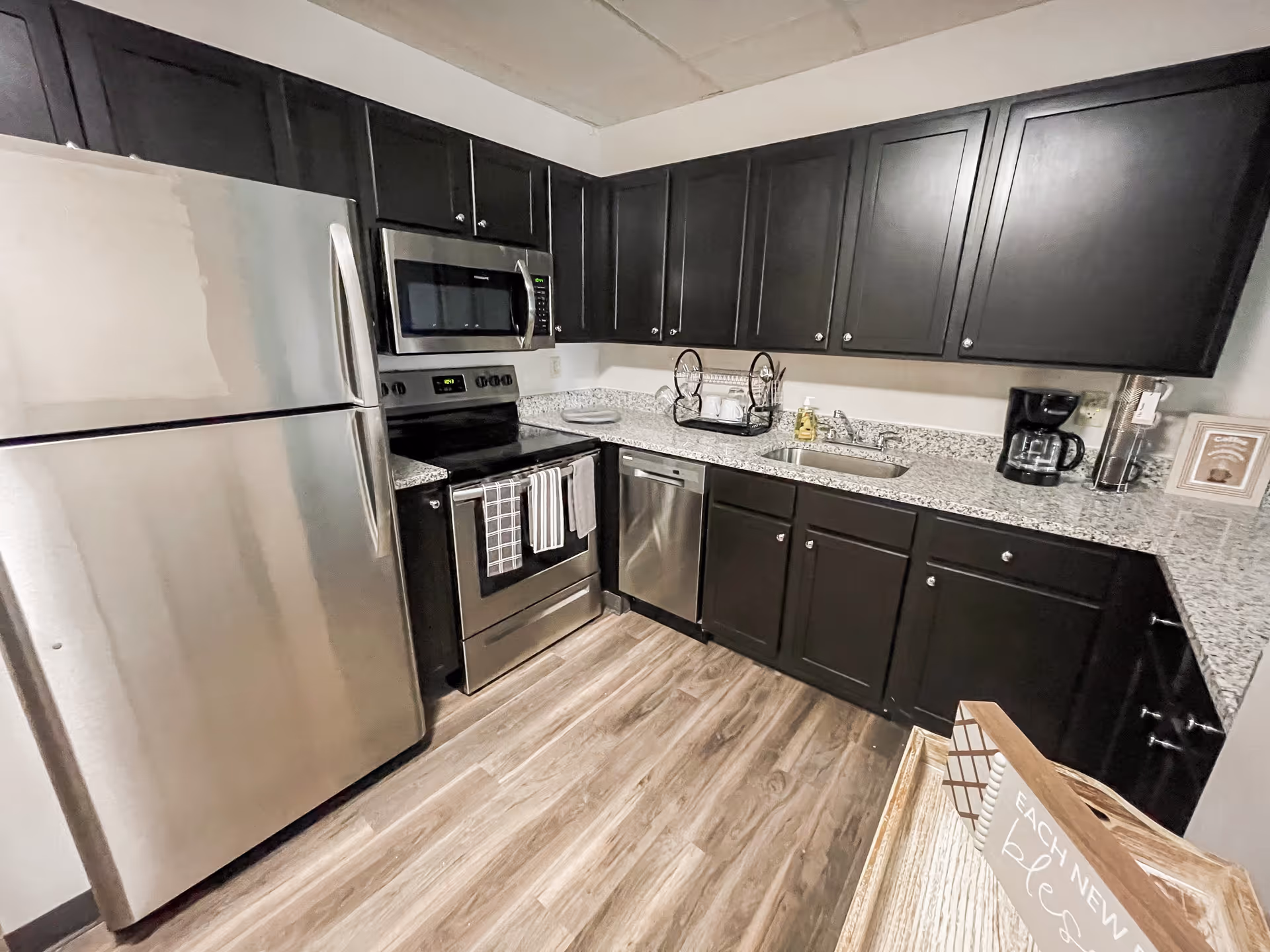 Modern kitchen with stainless steel refrigerator, stove, microwave, and dishwasher. Dark wood cabinets line the walls above and below a granite countertop with a sink, dish rack, coffee maker, and soap dispenser. The floor is wood-patterned, and a wooden tray with a sign reading 'Each new day is a blessing' is visible in the foreground.