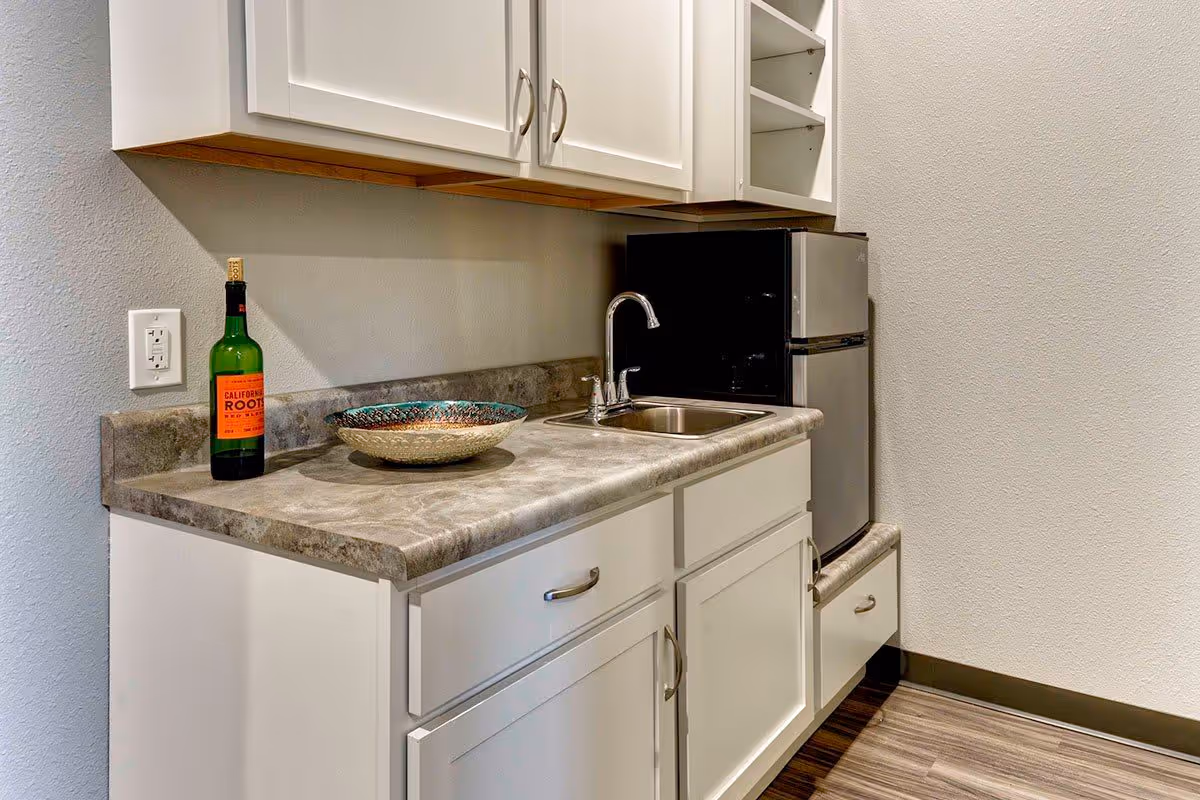 A small kitchenette area with white cabinets, a gray countertop, a stainless steel sink with a faucet, a small black and silver refrigerator, a decorative bowl, and a bottle of California Root wine on the counter.