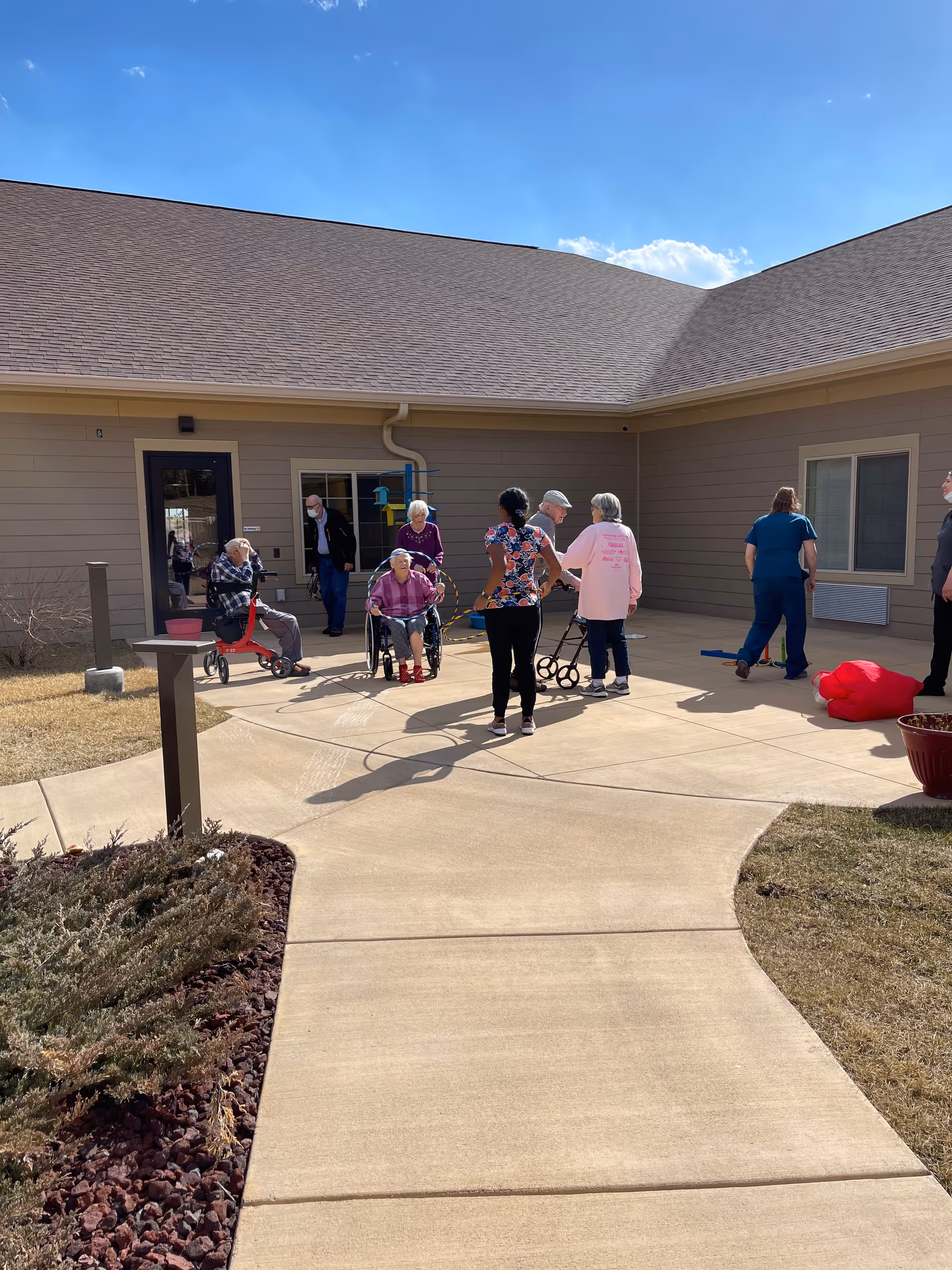Residents and staff gather in an assisted living courtyard with walkers and wheelchairs in front of a single-story building under a clear sky.