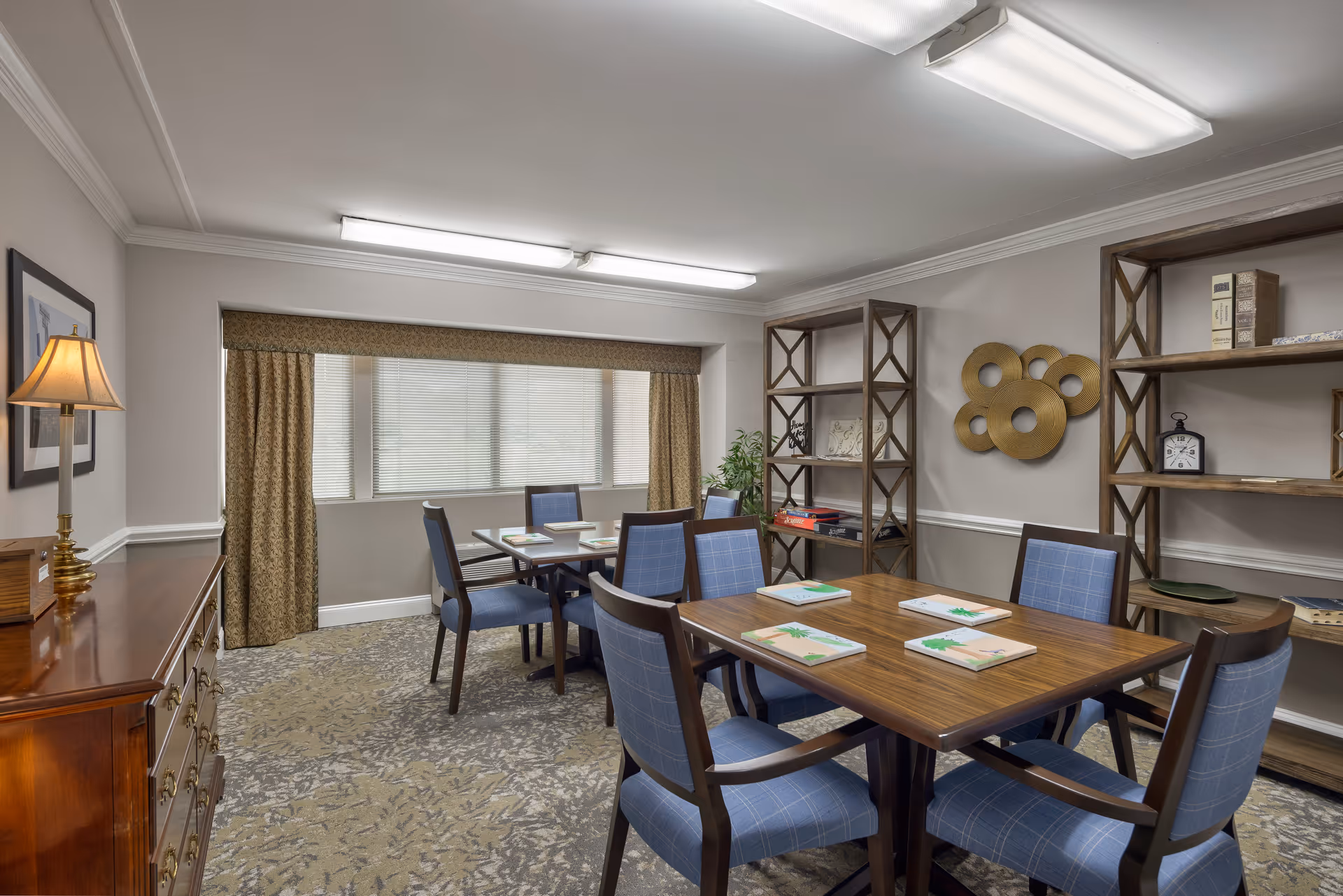 Well-lit communal dining/activity room with wooden tables, blue upholstered chairs, shelving, and a window with drapes.