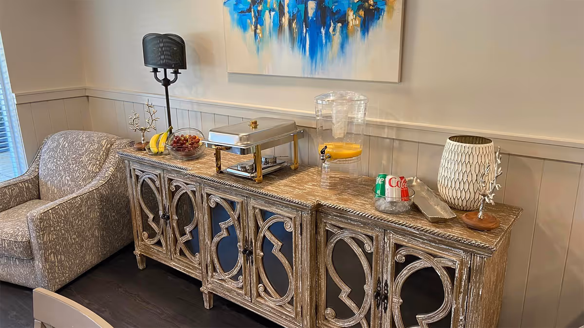 A cozy corner of a room featuring a patterned armchair next to a rustic wooden sideboard with decorative carved doors. On top of the sideboard are a lamp, a bowl of fruit, a covered serving dish, a beverage dispenser with orange juice, two soda cans in a metal holder, a decorative vase, and a small tree-shaped ornament. A colorful abstract painting hangs on the wall above the sideboard.