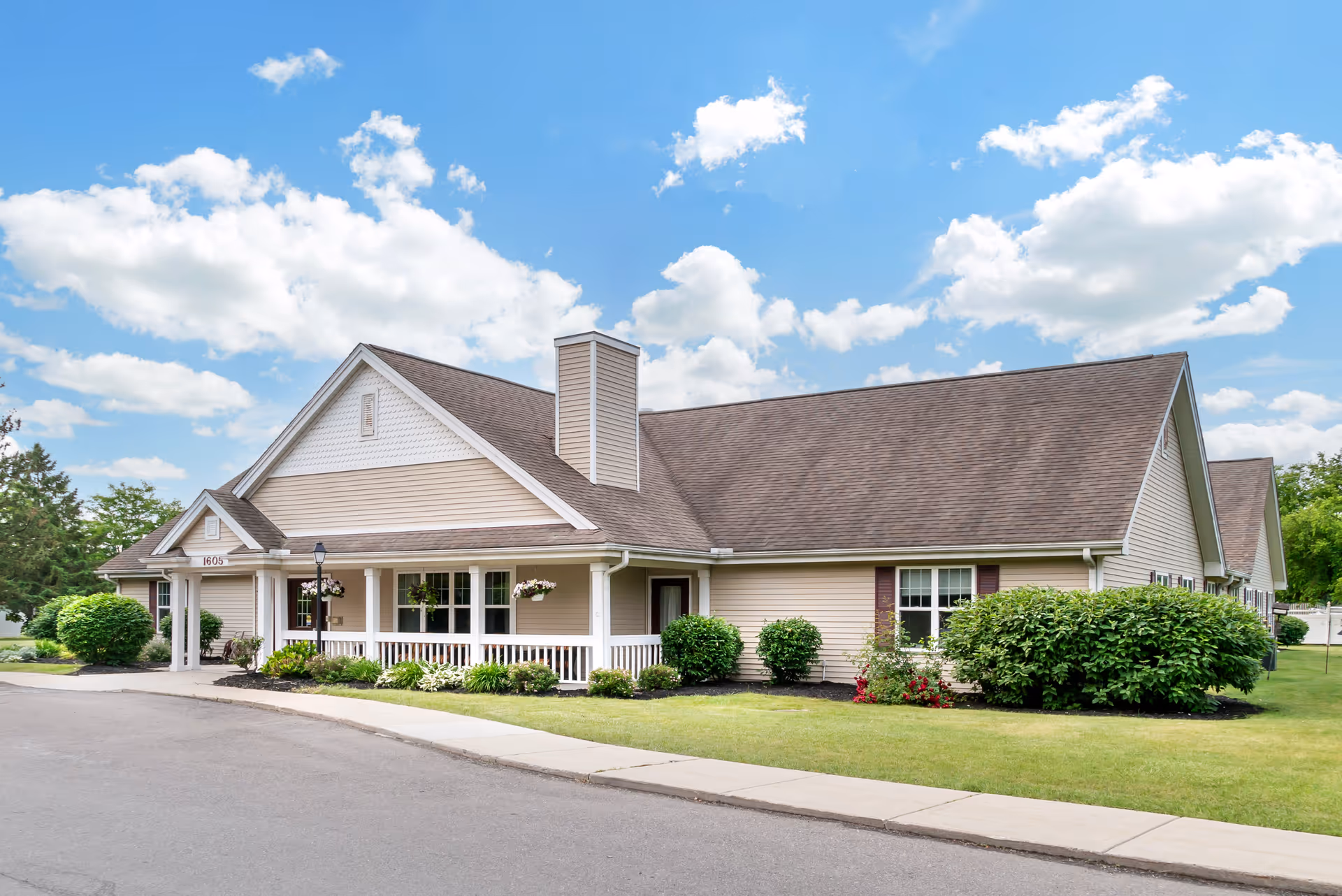 Exterior view of a single-story beige building with a brown shingled roof, white trim, and a small covered porch with white railings. The building is surrounded by neatly trimmed bushes and a well-maintained lawn under a partly cloudy blue sky.